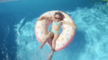 Yüzme havuzunda yüzen donutun üzerine su sıçratan mutlu küçük kız. Yüzerken ve havuzda oynarken güneş gözlüğü takan tatlı bir kız. Yaz tatilinde güneşli bir gün için eğlenceli bir aktivite