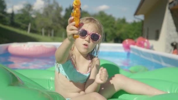 小女孩坐在漂浮的水面上 水枪飞溅在镜头前 嬉闹的小女孩漂浮在后院的游泳池里 直接对着相机喷水 — 图库视频影像