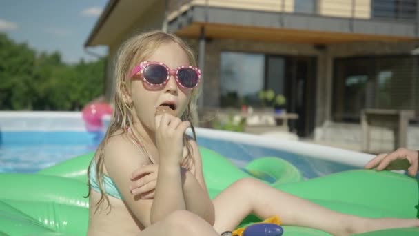 一个炎热的夏天 可爱的女孩在游泳池里飘浮 吃着冰激凌 可爱的小女孩在后院花园游泳池里享受巧克力冰淇淋吧 炎炎夏日的清爽茶点 — 图库视频影像