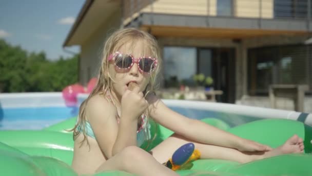 可爱的小女孩在游泳池里吃着冰激凌 向镜头挥手 可爱的女孩在后院花园游泳池里享用巧克力冰淇淋吧 炎炎夏日的清爽茶点 — 图库视频影像