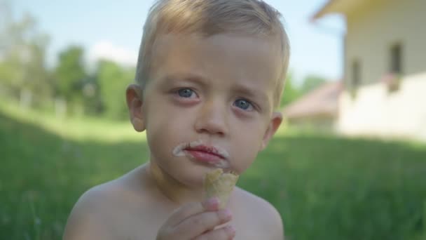 閉じる かわいい男の子は夏の日に庭の影でアイスクリームコーンを食べています 裏庭の日陰の部分でバニラアイスコーンを楽しんでいる愛らしい幼児 暑い夏の日の完全なリフレッシュ — ストック動画