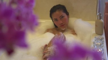 Genç Asyalı bayan rahatlatıcı bir köpük banyosu yapıyor ve kameraya bakıyor. Güzel Filipinli kadın ev banyosunda kaplıca tedavisinden zevk alıyor. Bayan kendini köpük banyosuyla şımartıyor..