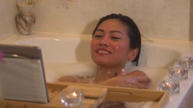 Güzel bir bayan, video sohbeti yaparken köpük banyosunda uzanıyor ve dinleniyor. Genç Filipinli kadın rahatlatıcı bir ortam içinde köpüklü bir banyo yaparken arkadaşıyla çevrimiçi video görüşmesi yapıyor..