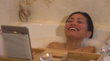 Neşeli Asyalı kadın köpük banyosu yaparken video aracılığıyla konuşuyor. Gülümseyen Filipinli kadın rahatlatıcı bir atmosferde köpüklü bir banyo yaparken arkadaşıyla görüntülü sohbet ediyor..