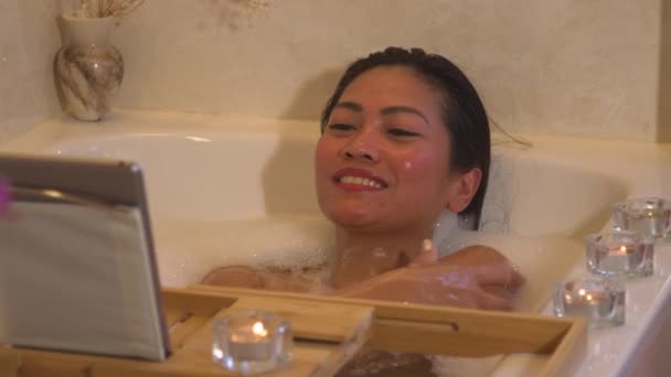 閉じる ビデオチャットをしながら きれいな女性が横になり バブルバスでリラックスしています 若いフィリピンの女性はリラックスした雰囲気の中で泡風呂を取りながら彼女の友人とオンラインビデオ通話を持っています — ストック動画