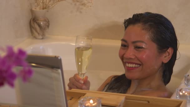 バブルバスの中で若い女性の笑顔は ビデオ通話を介してロマンチックなデートを持っています 美しいですフィリピン人女性飲料ガラスのシャンパンで泡でいっぱいながら ビデオ通話を介して友人とチャット — ストック動画