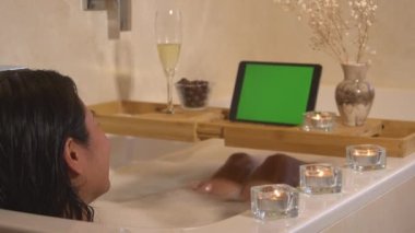 Genç bayan köpük banyosunda dinleniyor ve çevrimiçi video görüşmesi ile sohbet ediyor. Güzel siyah saçlı kadın banyoda küvette uzanıyor ve iyi bir arkadaşla dijital tablette görüntülü sohbet ediyor..