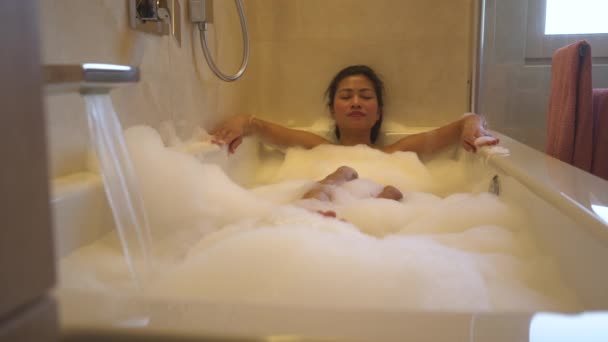 蛇口から水が流れている間 泡でいっぱいのお風呂でリラックスしているきれいな女性 美しい若いアジアの女性は 自宅のバスルームで泡風呂に横たわっている間 スパとリラックスして自分自身を治療 — ストック動画