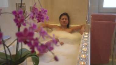 Güzel, çiçekli bir orkide ve güzel, genç bir bayan köpük banyosunda dinleniyor. Genç ve güzel bir kadın küvette eğleniyor ve dinleniyor. Spa tedavisi için sakin ve keyifli bir atmosfer..