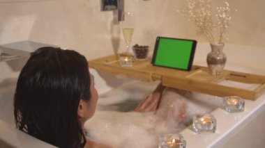 Güzel bayan köpük banyosunda oturuyor ve online video aracılığıyla sohbet ediyor. Güzel siyah saçlı kadın dijital tablette görüntülü konuşma yapıyor. Uzun ve yoğun bir günün sonunda rahatlatıcı anlar.
