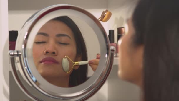 年轻女人用面部辊自我按摩她美丽的脸庞 漂亮的菲律宾女士在年轻的面部治疗 镜像中的女性在照顾自己的容貌 — 图库视频影像