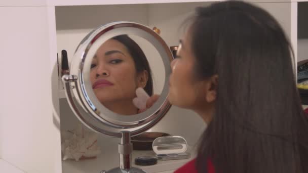 年轻的女士用玉石刮胡刀按摩自己的脸 用于面部整容 美容美发亚洲女子在用瓜沙刮胡刀进行抗衰老推拿的化妆品镜中的反映 — 图库视频影像