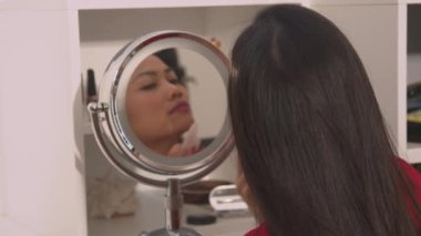 Gua sha kazıyıcı kullanarak kozmetik aynada güzel bir kadının yansıması. Filipinli genç bayan cilt sertliği ve yüz gerdirme masajı için yeşillik silindiri kullanıyor..