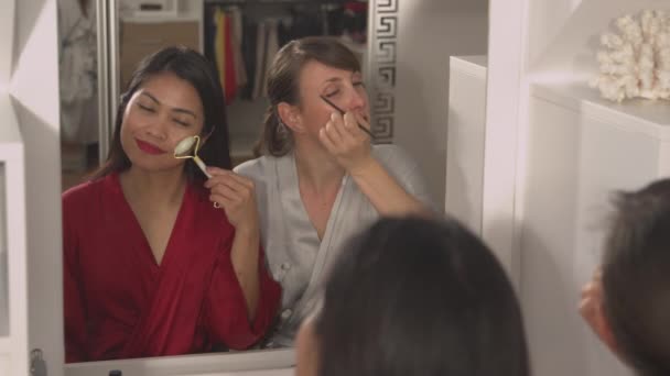 閉じる 鏡の反射に構成を適用魅力的な若い女性のビュー アジア人と白人の女性は美容製品を使用して夜の外出の準備をします 美の治療を受けている二人の女性 — ストック動画