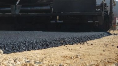 Paver makinelerinin arkasından yeni bırakılmış asfaltı daha yakından görebiliriz. Ağır iş makineleri, sabah güneşiyle yola dökülüyor ve yeni asfalt dökülüyor..