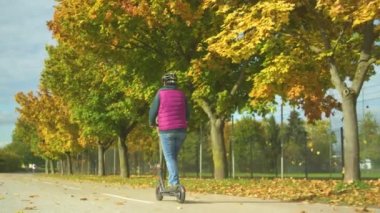 Sonbahar ağaçlarının yanında elektrikli scooter kullanan yaşlı bir kadının arka görüntüsü. Kıdemli bayan, modern çevre dostu bir yol kullanıyor. Kasabada e-scooter sürmek için güzel bir sonbahar günü..