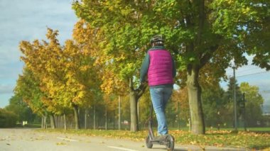 Kıdemli bayan modern ve çevre dostu ulaşım için e-scooter kullanıyor. Sonbahar ağaçlarının yanında elektrikli scooter kullanan yaşlı bir kadının arka görüntüsü. Scooter sürmek için güzel bir sonbahar günü..