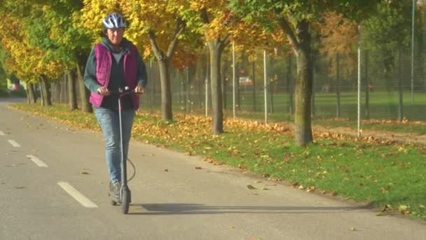 上了年纪的妇女骑着电动车沿着秋树林荫大道骑自行车 老太太喜欢在一个美丽的秋天骑电动车 现代生态友好型交通方式 — 图库视频影像