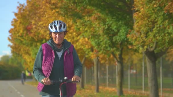 閉じる 陽気シニア女性は秋の日にE Scootlerで運転を楽しんでいます 秋のツリーライン通りに沿ってサイクルパス上の電動スクーターに乗る高齢女性 スクーターに乗るための素敵な秋の日 — ストック動画
