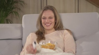 Evde komedi filmi izlerken cips yiyen ve gülen bir kadın. Güzel bayan televizyon izlerken evde oturma odasında eğleniyor. Kış akşamları dinlenme etkinliğinde olan genç bir kadın..