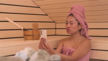 Neşeli, genç Asyalı bayanın Fin saunasında rahatlamasını ve selfie çekmesini sağla. Sosyal medya içeriği oluşturmak için sağlık tedavisinde akıllı telefon kullanarak pembe havluya sarılmış güzel bir kadın etkili..