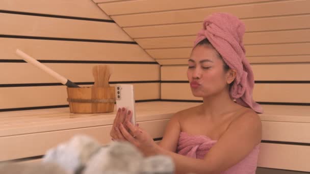 在芬兰桑拿浴室休息一下 并进行自我锻炼 与快乐的年轻亚洲女士亲密接触 漂亮的女性影响者用粉红毛巾包裹着 用智能手机在健康治疗中创造社交媒体内容 — 图库视频影像