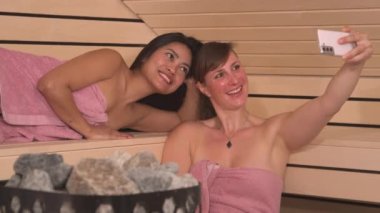 Mutlu genç kadınlar selfie çekiyor ve spa ziyaretinden hatıralar yaratıyor. Pembe havluya sarılı güzel kız arkadaşlar ahşap Finlandiya saunasında sağlık hizmetinde eğleniyor ve eğleniyorlar..