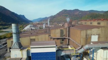 Arka planda sonbahar manzarası olan endüstriyel binaların üzerinde uçmak. Sonbahar mevsiminin renkli gölgeleriyle çevrili şehir ve dağlarla çevrili pitoresk alp vadisindeki çelik fabrikası..