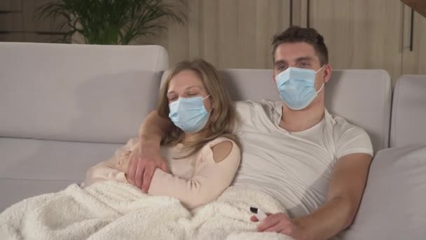亲密接触 夫妇在病假期间戴着防护面罩看电视 秋寒和流感四处蔓延 年轻夫妇躺在舒适的沙发上 躺在毯子下 得了季节性流感 — 图库视频影像