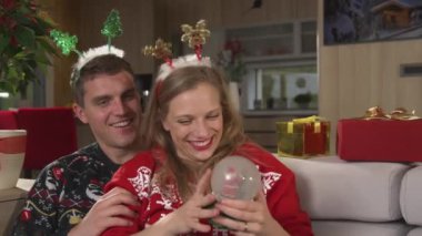 Kocasının kollarındaki neşeli, neşeli kadın bir kar kubbesiyle eğleniyor. Noel tatilini kutlayan ve evde birlikte rahatlayan genç bir çift..