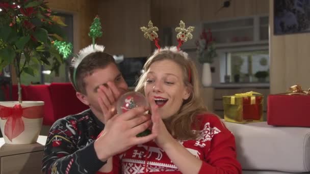 亲密接触 快乐的男人用一个圣诞雪球给他美丽的女士带来惊喜 年轻夫妇欢欢喜喜地庆祝圣诞佳节 并在家中客厅里共度轻松的时光 — 图库视频影像