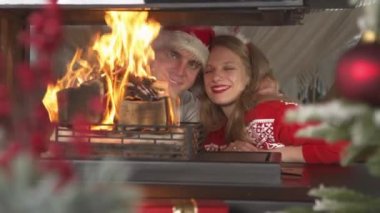 Cute couple in love showing affection while watching burning fire in fireplace. Cheerful man and woman wearing Christmas sweaters and enjoying festive ambiance in home living room on Christmas Eve.
