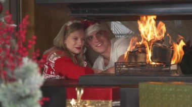 Loving Christmas couple leaning on each other and looking at burning fireplace. Cheerful man and woman wearing festive sweaters and enjoying romantic ambiance in home living room on Christmas Eve.