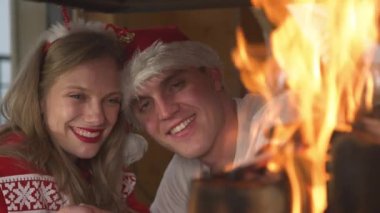 CLOSE UP: Romantic couple leaning on each other and watching burning fireplace. Smiling man and woman wearing festive sweaters and enjoying romantic ambiance in home living room on Christmas Evening.