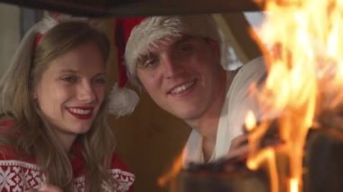 CLOSE UP: Smiling man and woman in festive mood looking at burning fireplace. Cheerful young couple wearing festive sweaters and enjoying romantic ambiance in home living room on Christmas Evening.