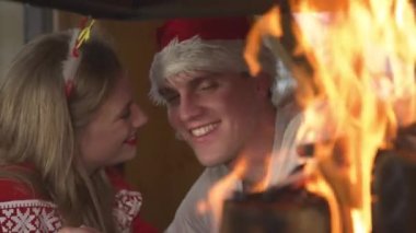 CLOSE UP: Lovely couple share a kiss on Christmas while looking at fireplace. Cheerful young couple wearing Christmas sweaters and enjoying romantic ambiance in home living room on festive Evening.