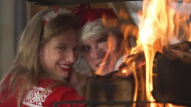 CLOSE UP: Burning fire flames and cute romantic couple leaning on each other. View of fireplace and young twosome in love enjoying romantic atmosphere in home living room on festive winter Evening.