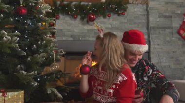 Smiling romantic couple decorating Christmas tree with red baubles at fireplace. Young man and woman preparing Christmas decoration in their home living room for celebrating festive winter holidays.