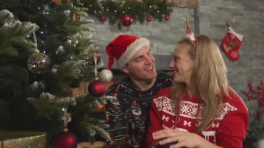 Pretty couple decorating Christmas tree when man surprises his lady with a gift. Young man and woman wearing festive outfit celebrating Christmas holiday at beautifully decorated home living room.