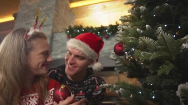 CLOSE UP: Beautiful woman hangs red bauble on Christmas tree and kisses her man. Lovely young couple wearing festive outfit celebrating Christmas holiday at beautifully decorated home living room.