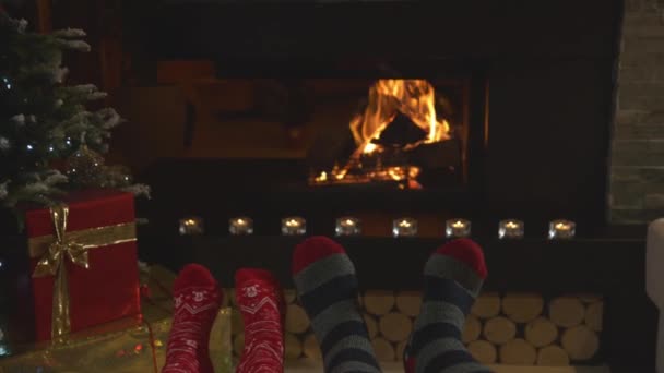 閉じる クリスマスの靴下に足を揺らし 暖炉で暖まるカップル クリスマスの夜に美しく装飾された自宅のリビングルームの快適さで男と女を楽しんでリラックス — ストック動画