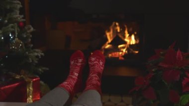 CLOSE UP: Wiggling female feet in Christmas socks warming up by the fireplace. Woman wearing red socks with festive pattern and relaxing at fireside, decorated Christmas tree and blooming poinsettia.