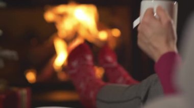 CLOSE UP Lady drinking hot tea while relaxing by fireplace and Christmas tree. Female person enjoying and resting in warm relaxing ambiance of her festively decorated home living room on Christmas Eve