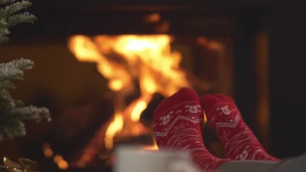 看到圣诞袜子的摆动的脚 后面有壁炉 在温暖舒适的客厅里 穿着红色袜子 头戴喜庆冬季图案 在燃烧的炉火前休息的女人 — 图库视频影像