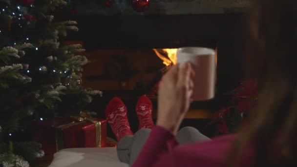 在壁炉边和圣诞树旁 年轻女子放松的背影 圣诞前夕 穿着印有节日图案的红袜 边喝茶边在炉边休息的女性 — 图库视频影像