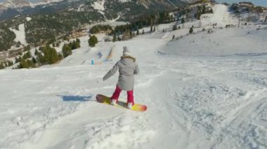 Young female learning to ride a snowboard and making turns on snowy ski slope. Girl snowboarder lands in snow after losing balance while snowboarding down the piste at snow-covered mountain ski resort