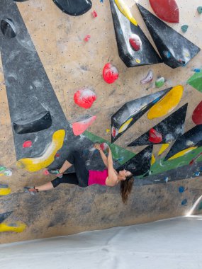 Young Caucasian woman gets inverted while climbing indoors at a colorful training hall. Experienced female rock climber climbs a challenging artificial climbing wall while training during the winter.