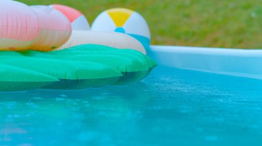 CLOSE UP, DOF: Cinematic shot of a backyard pool filled with floaties getting caught in an autumn rainstorm. Crystal clear rain droplets fall into the empty turquoise water and colorful floaties.