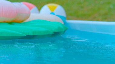 CLOSE UP, DOF: Crystal clear rain droplets fall into the empty turquoise water and colorful floaties. Cinematic shot of a backyard pool filled with floaties getting caught in an autumn rainstorm.