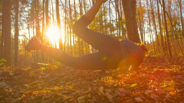 LENS FLARE, LOW ANGLE: Unrecognizable female jogger stumbles to ground while running in forest. Fit young woman running through the fall colored woods at sunset trips and falls into heap of leaves.